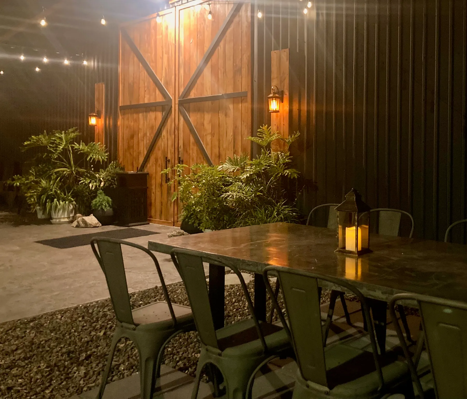 The patio after dark: the barn doors lit warm, string lights overhead, and a lantern glowing on the table