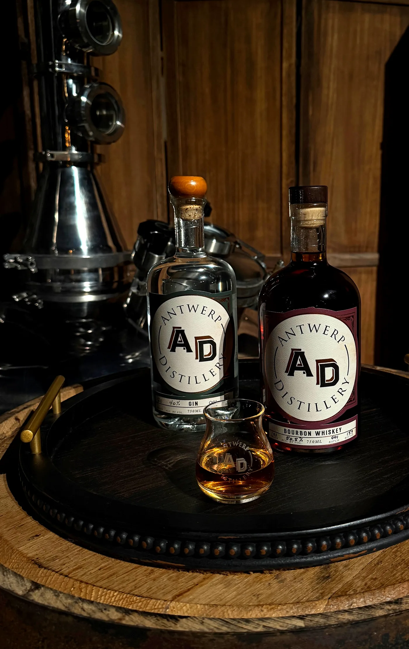 Antwerp Distillery gin and bourbon whiskey bottles with a tasting glass on a barrel head, stills behind