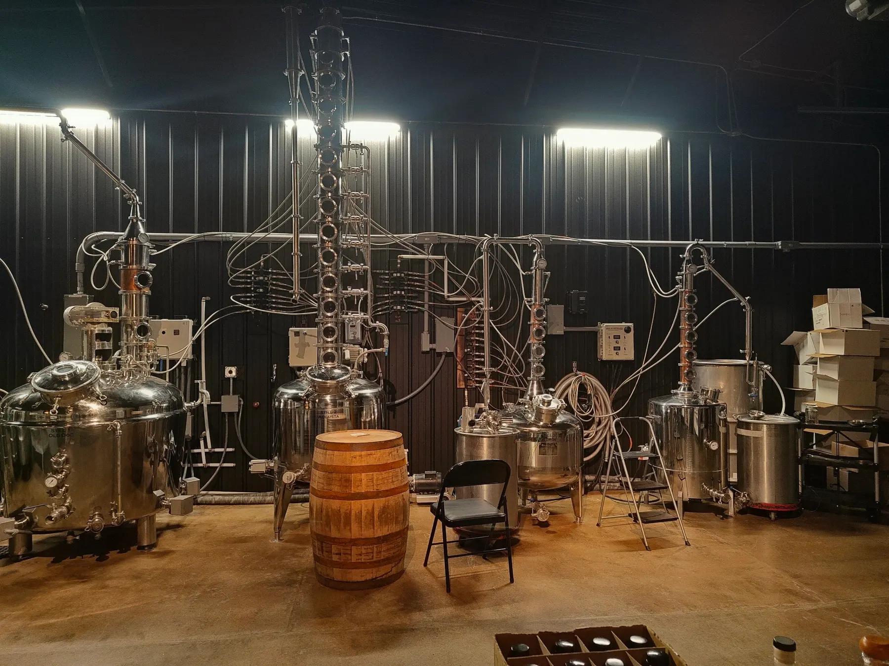 The full row of six stills on the Antwerp Distillery stillhouse floor, with a bourbon barrel in the foreground